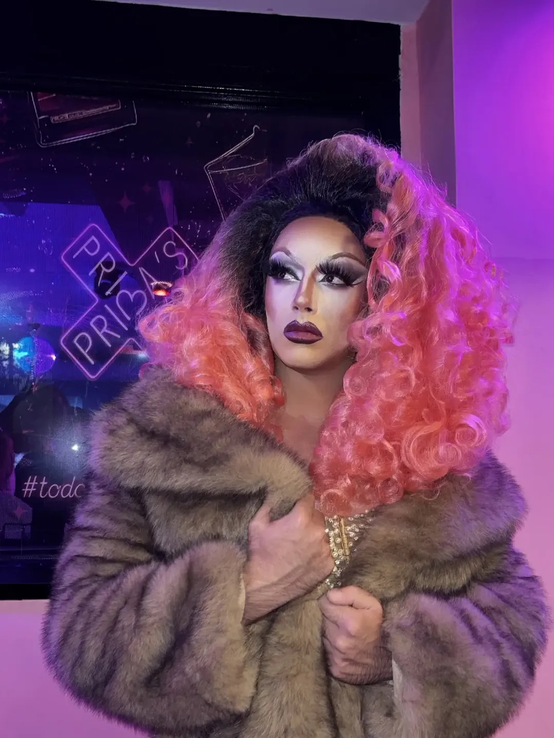Una drag con peluca rosa y abrigo de pelo delante del neón de Prima’s